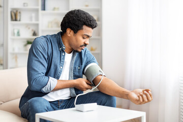 A focused young African American man sits comfortably on a couch while he wraps a blood pressure...