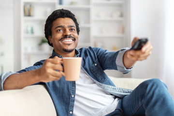 A cheerful African American man is lounging comfortably on a sofa with a warm beverage in one hand...