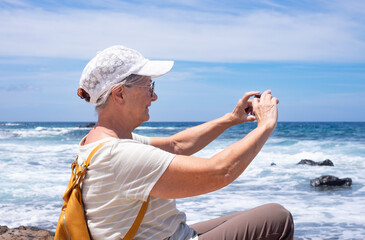 Smiling mature senior woman with backpack and cap admiring the sea waves taking a photo for memory....