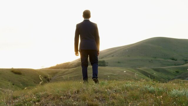 A man in a black business suit has a glow around his silhouette. A man with a tired gait stops and falls to his knees. A man looks at the horizon, where the sun is hidden.
