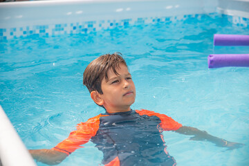 Boy in swimwear floating in a blue pool, looking contemplative under a clear sky