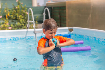 Boy in a home pool playing with a ball, a blue swim noodle floating in the background, garden...