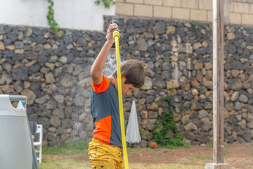 Viewed from behind, a child exerts power over a garden hose, spraying water upwards, creating a...