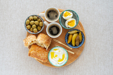 Israeli style breakfast including baked pastry Bourekas, green olives, pickled peppers, yoghurt, boiled eggs, cups of coffee on wooden tray.