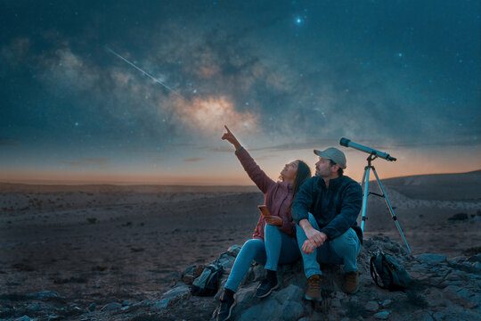 two people sitting in the desert watching the stars and Milky Way next to a telescope, stargazing and exploration concept 
