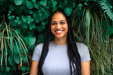 young smiling black female with afro braids is staying near a wall covered with green leaves
