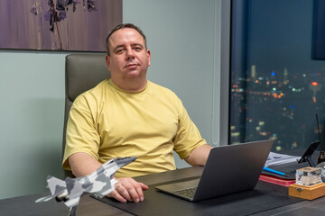 Overweight confident man is sitting at desk with laptop and toy airplane in modern office by window...