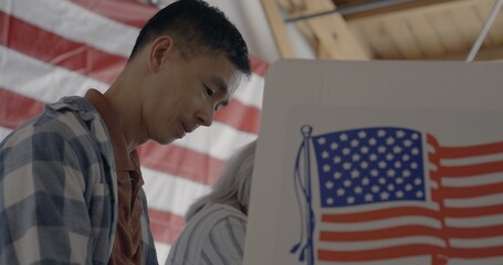 Low viewpoint, profile, looking up at Asian man voting at booth while considering candidates in...