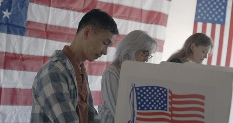 Profile view across voting booths with focus on Asian man casting votes in foreground. Moves from medium shot to close up. Senior woman and young woman voters behind, with US flag in background. 4K DC
