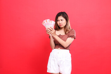 Expression of a young Asian woman, sad, candid, both hands holding rupiah currency, wearing a brown...
