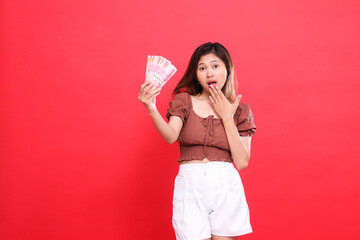 the expression of a young Asian woman shocked at the camera, covering her mouth and holding rupiah...