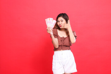 gesture of a cheerful young Asian girl holding rupiah money in front and a telephone sign. I am...