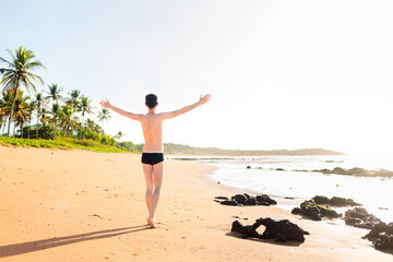 Brazilian man wearing swimwear, with his arms raised to express gratitude and happiness for enjoying summer vacation on a paradise beach