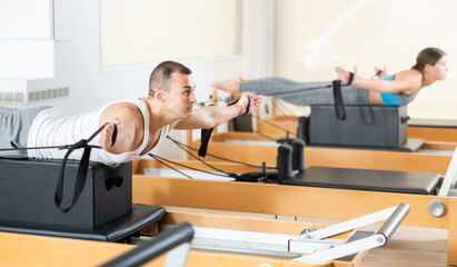 Concentrated man in comfortable sportswear performing series of exercises with straps on Pilates reformer during rehabilitation workout in fitness studio..