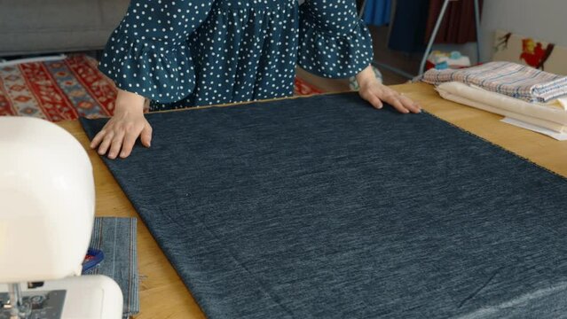 An overhead view of a woman in a polka dot blouse preparing denim fabric on a table next to a sewing machine, with various crafting materials in the background.