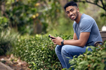 Fototapeta premium Phone, Indian man and student in park, outdoor and typing in app with connection to social media and portrait. University, education and person in campus with mobile, texting and chat for update