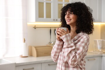 Beautiful young woman in stylish pyjama with cup of drink in kitchen, space for text