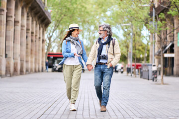 Happy adult Caucasian tourist couple looking at each other walking holding hand for street in city. Mature people in love enjoying romantic getaway. Positive relationships and spring retiree holidays