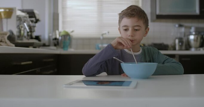A young boy is seated at a modern kitchen table, focused on a digital tablet with a bowl of cereal in front of him.