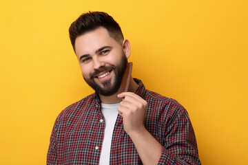 Handsome young man combing beard on yellow background
