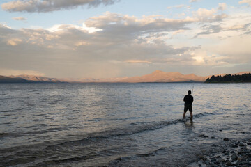 Naklejka premium Sports and recreation. View of a man fishing from the rocky shore in Nahuel Huapi lake at sunset.