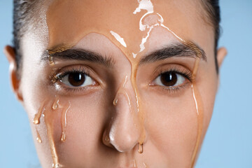 Beautiful young woman with dripping honey on her face against blue background, closeup