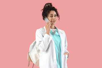 Female African-American medical intern with backpack talking by mobile phone on pink background