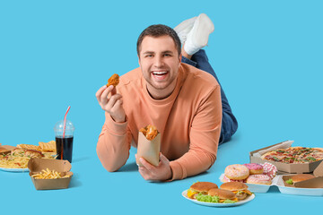 Young man with unhealthy food lying on blue background. Overeating concept