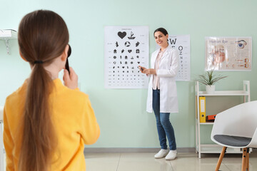 Female ophthalmologist checking little girl's eyesight near test chart in clinic