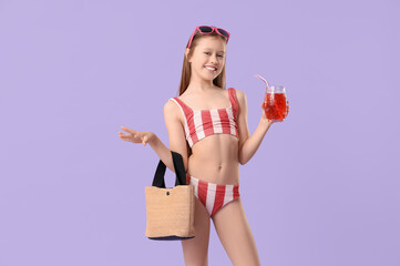 Cute little girl in swimsuit with beach bag and glass of lemonade on purple background