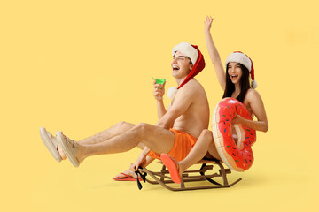 Young couple in Santa hats with cocktail and swim ring on sledge against yellow background....