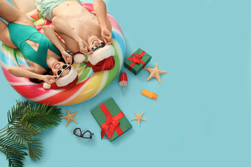 Young couple in Santa hats with beach accessories lying on blue background, top view. Christmas in July