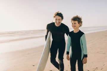 Father and son in wetsuits walking on the beach with a surfboard