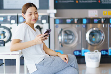 Asian woman using smartphone while waiting her clothes washed at laundry shop.