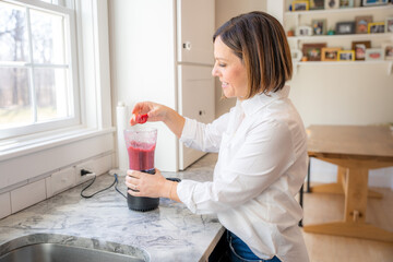 Cute middle age woman mixes up a berry smoothie in a blender in a white kitchen