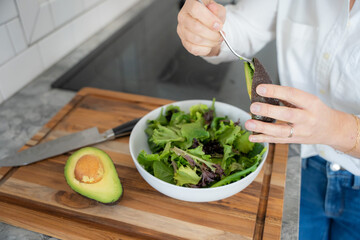 Cute middle aged woman adds avocado to a mixed greens salad in her white kitchen
