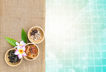 Thai dry herbs in round wooden bowl with Plumeria flower on hessian fabric background, spa concept, Dry Bael fruit and tea with Thai spice in wooden cup with space on swimming pool water background, w