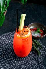 Bloody Mary cocktail with celery, hot sauce, lemon and ice in a glass on a dark background. Spicy...
