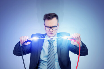 A man in a dark suit and glasses holds two thick wires with clips between which a powerful...