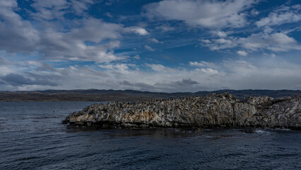 Many cormorants have settled on a rocky island in the Beagle Channel. Ripples on the blue ocean water. Clouds in the azure sky. Mountains in the distance. Argentina. Tierra del Fuego Archipelago.