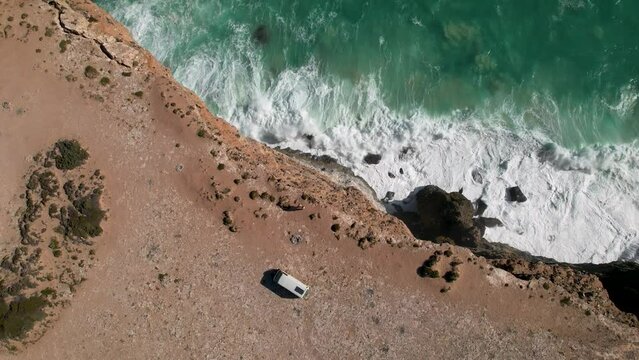 Birdseye view of a couple standing near a camper van on a cliff edge with the waves crashing against the rocks below. This is Bunda Cliffs, a campsite near the famous Nullarbor in South Australia.