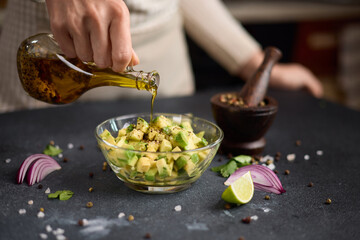 Woman pours olive oil onto Chopped ingredients in wooden bowl - avocado, onion and pepper