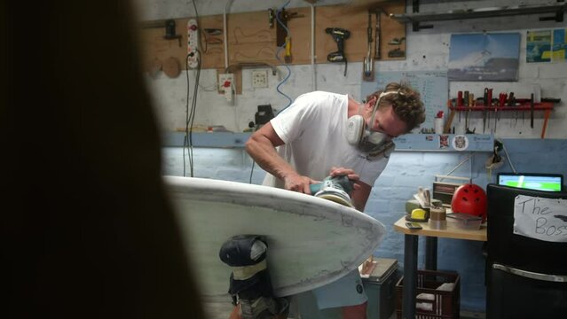 Entrepreneur surf board maker using sanding power tool to shape white board in workshop with protective gear - slow motion