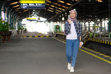 happy asian muslim woman wearing casual outfit walking on train station platform. traveling concept