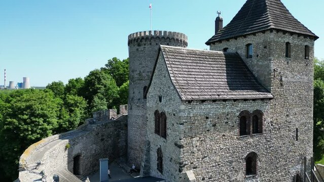 Medieval Bedzin castle with a turret, stone walls, and courtyard during a beautiful summer day surrounded by lush greenery, under a clear blue sky.