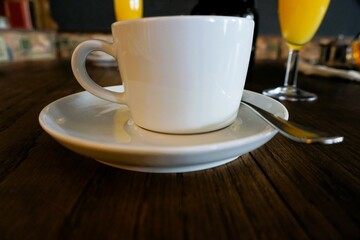 Weiße Tasse Kaffee mit Untertasse auf braunem Holztisch mit Sektgläsern gefüllt mit Orangensaft...
