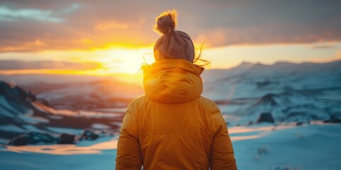 back view image of an unrecognizable woman in a yellow jacket standing and observing a sunset in a snowy mountain landscape