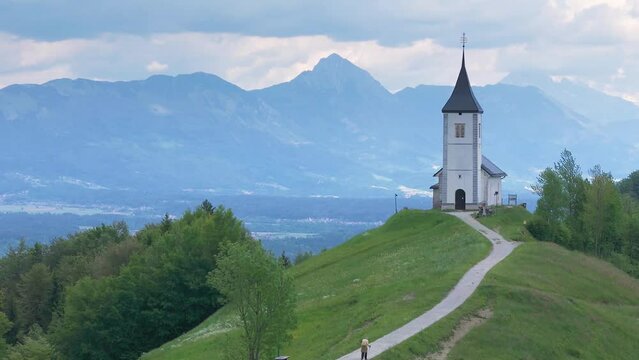 Flying by the Catholic church at the top of the hill, the St. Primus and St. Felician church at Jamnik, Slovenia, Kamnik Savinja Alps in background. Drone shot, 4K, 60FPS