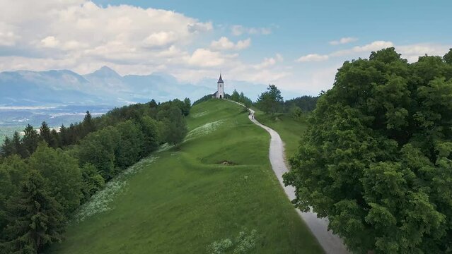 Flying towards Catholic church at the top of the hill, the St. Primus and St. Felician church at Jamnik, Slovenia, Alps in background. Tourism, hiking, pilgrimage concepts. Drone shot, 4K, 60FPS