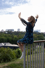 elegant woman in a dress sits on the railing with her hands raised to the sky. Fantasies, dreams, aspirations for a bright future